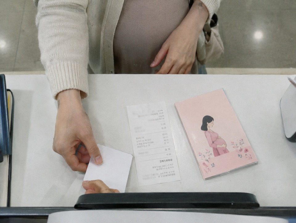 병원 접수대에서 카드를 건네는 임산부의 손과 산모수첩